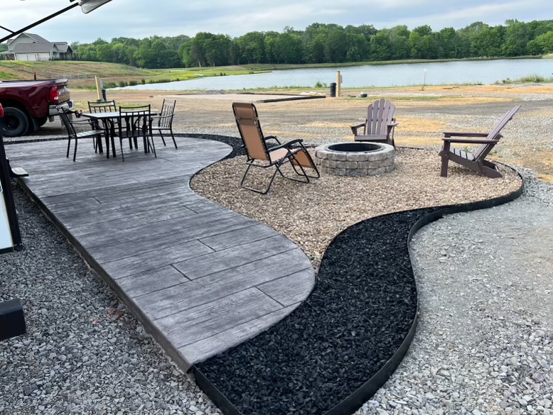A landscaped patio area with a table and chairs on stamped concrete, lounge chairs around a stone fire pit on gravel, bordered by black mulch, with a lake and trees in the background.
