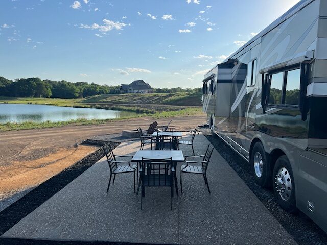 A large RV is parked beside a patio with tables and chairs, overlooking a pond and open grassy landscape under a blue sky with scattered clouds. A house is visible in the distance.