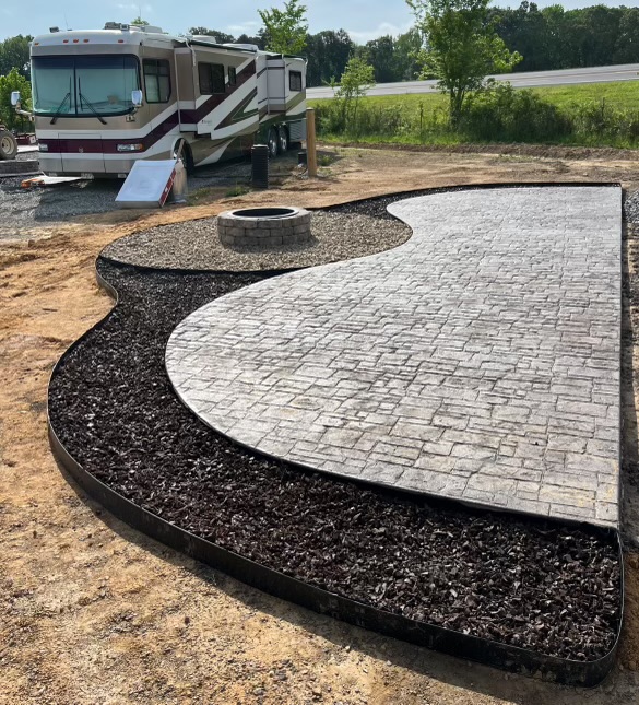 A paved patio area with a fire pit, bordered by dark mulch and light gravel, next to a motorhome parked on a dirt lot with trees and a road in the background.