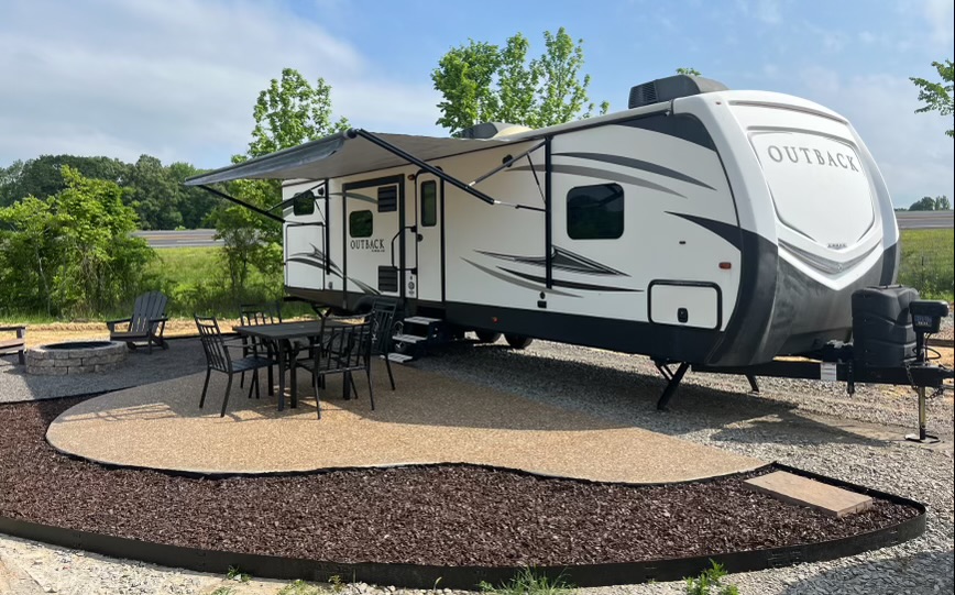A white and gray Outback travel trailer is parked on a gravel lot with an awning extended. Nearby, there is a patio area with a table, chairs, Adirondack chairs, and a fire pit, surrounded by trees and greenery.