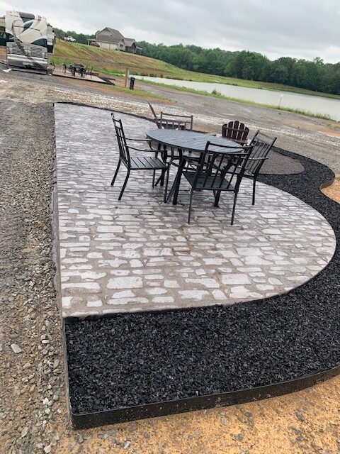 A patio with a metal table and four chairs sits on gray pavers next to black mulch. An Adirondack chair is nearby. In the background, there are RVs, grass, a pond, and trees under a cloudy sky.