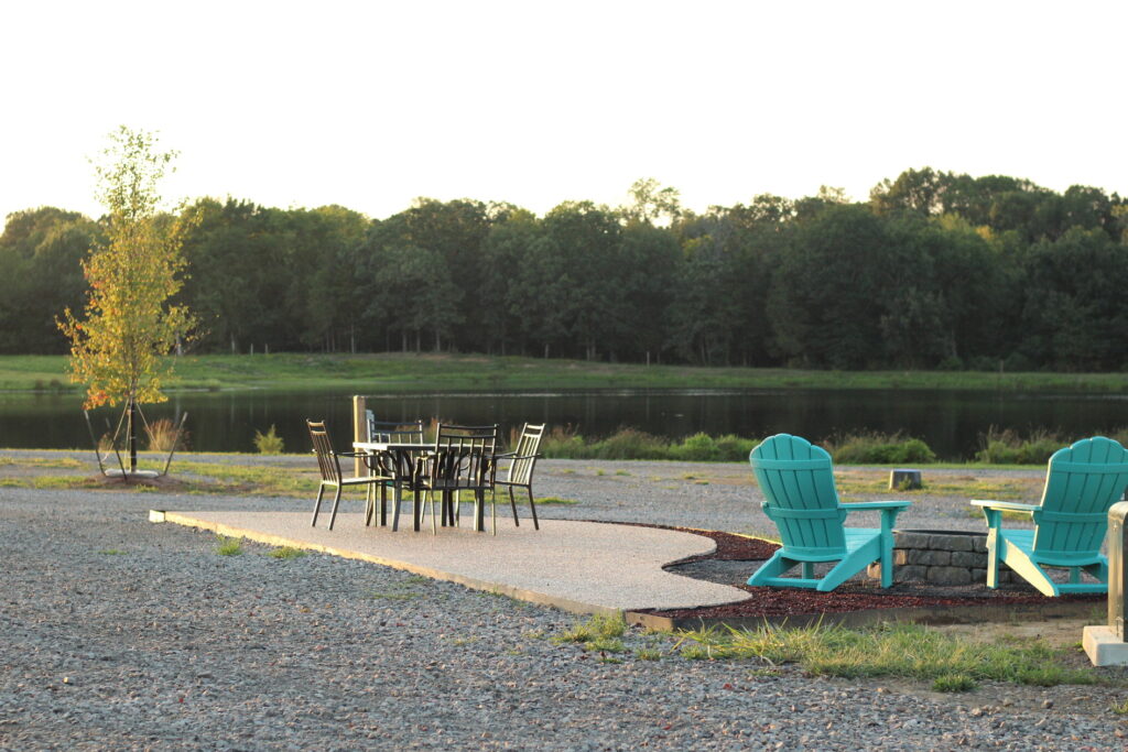 Outdoor seating area near a calm lake, featuring a table with black chairs and two turquoise Adirondack chairs by a fire pit, surrounded by gravel and trees in the background.
