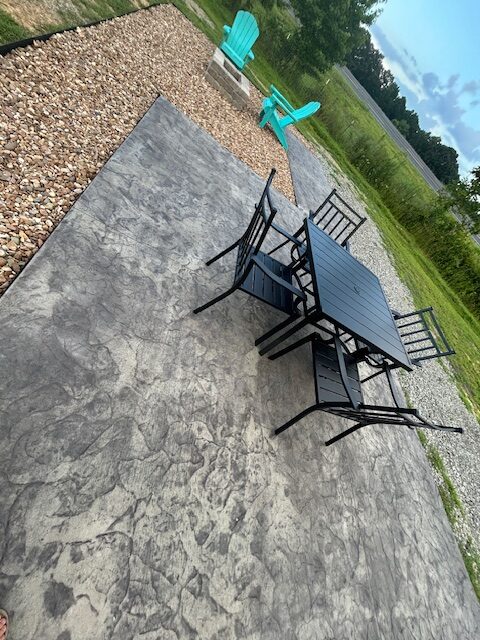 A patio with a black metal table and chairs on stamped concrete, bordered by gravel and rocks. Two turquoise Adirondack chairs sit near a fire pit in a grassy backyard under a partly cloudy sky.