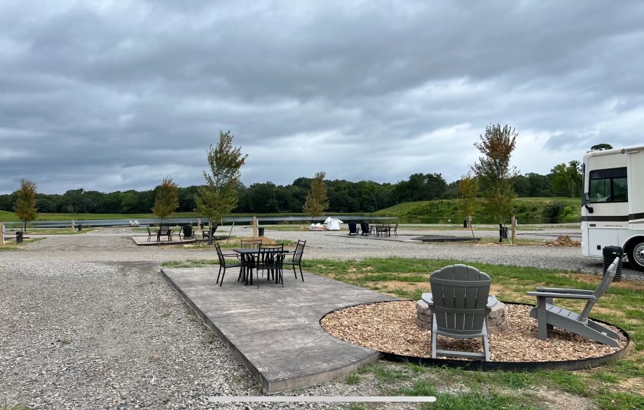 Outdoor RV campground with paved seating areas, tables, chairs, and fire pits. Some trees and grassy patches are visible, and an RV is parked on the right. A pond and wooded area are in the background under a cloudy sky.