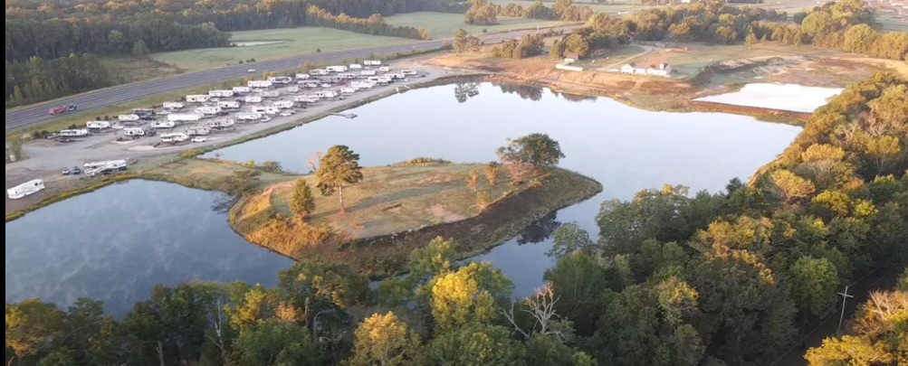 Aerial view of a pond with a small island, surrounded by trees and a nearby RV park, with a road and open fields in the background.