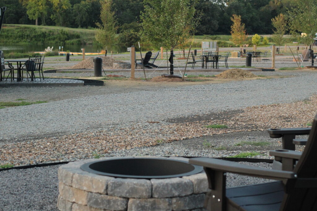 Outdoor seating area with gravel paths, fire pits, Adirondack chairs, tables, and chairs, set near a pond with trees in the background.