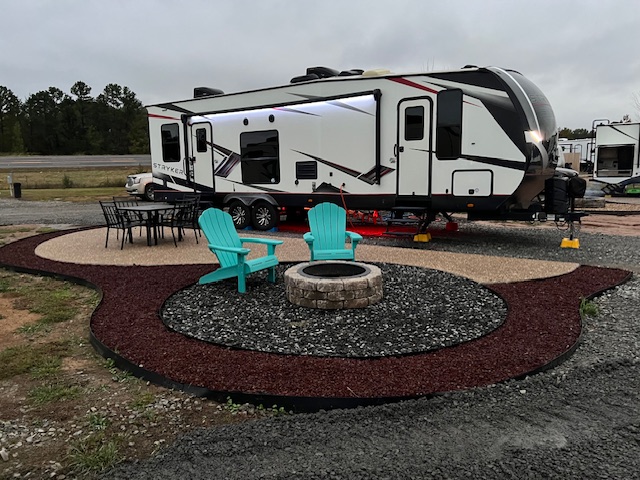 A travel trailer parked at a campsite with two teal chairs and a fire pit on a landscaped gravel and mulch patio, with a dining table set nearby.