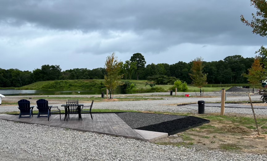 A gravel lot with a wooden patio features four blue chairs and a black table; trees and grassy area are visible in the background under a cloudy sky.