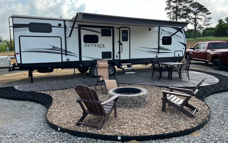 A travel trailer labeled "Outback" is parked beside a red truck. In front, there is a fire pit surrounded by Adirondack chairs, a lounge chair, and an outdoor dining set.