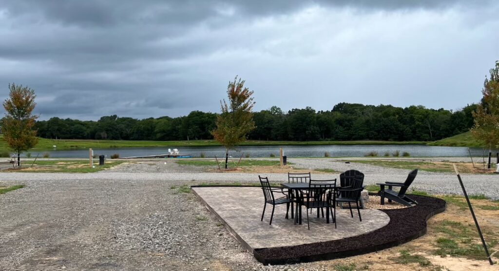A patio with outdoor tables and chairs overlooks a pond surrounded by grass and trees under a cloudy sky.